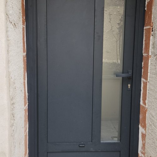 Porte d’entrée en aluminium, gris anthracite (RAL 7016) composée d’une allège en panneau plein en partie basse, d’un panneau plein vertical et d’une autre partie vitrée verticale avec vitrage dépoli.