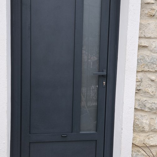 Porte d’entrée en aluminium, gris anthracite (RAL 7016) composée d’une imposte en partie haute avec vitrage dépoli, d’une allège en panneau plein en partie basse, d’un panneau plein vertical et d’une autre partie vitrée verticale avec vitrage dépoli.