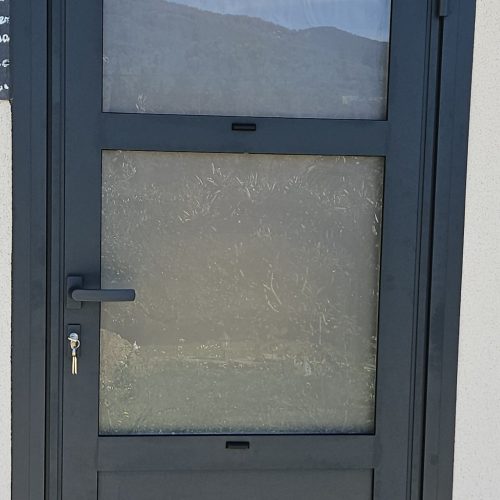 Porte d’entrée en aluminium, gris anthracite (RAL 7016) en 3 parties égales avec panneau plein en partie basse et deux vitrages dépolis en partie haute.
