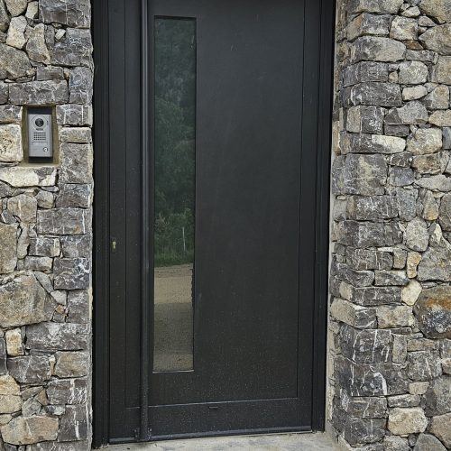 Porte d’entrée en aluminium, de couleur gris anthracite RAL 7016, équipée d’un bâton de maréchal côté extérieur et d’une poignée côté intérieur mais aussi d’une gâche électrique avec interphone